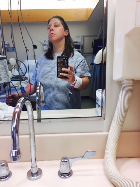 Image of a white woman in a hospital setting. She is pointing her phone at a mirror and scowling. 